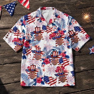 May include: White short-sleeved button-down shirt with a patriotic design. The shirt features a repeating pattern of stars, American flags, fireworks, and floral elements. The design includes a face wearing a patriotic hat. The shirt is on a wooden surface.
