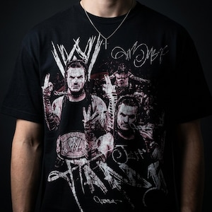 May include: A black short-sleeved t-shirt featuring a graphic print of professional wrestler Jeff Hardy. The design includes the WWE logo, multiple images of the wrestler, and stylized white text overlays. A thin silver chain with a cross pendant is visible.