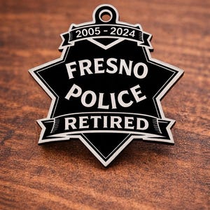May include: A black and white Fresno Police Retired badge with the years 2005-2024. The badge is in the shape of a star with the words "Fresno Police Retired" in white lettering. The badge is set against a wooden background.