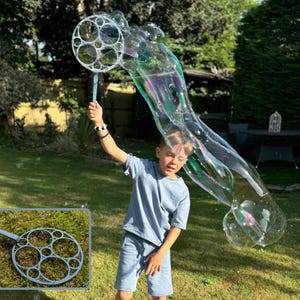 May include: A child holds a bubble wand, creating a large, iridescent bubble in a sunny outdoor setting. The wand has a circular design with multiple holes. The child is wearing a blue t-shirt and shorts. The background includes green grass and trees.