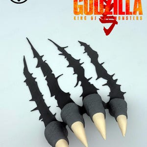 May include: A black claw-shaped wall decoration featuring three jagged, textured slashes with grey, scaled finger-like bases and pointed, off-white tips. The text GODZILLA KING OF THE MONSTERS and a Japanese character are printed in red and orange.
