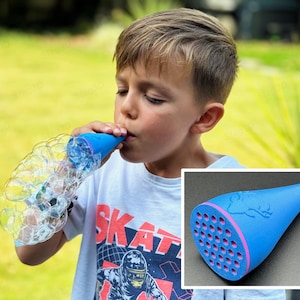 Puede incluir: Primer plano de una varita de burbujas azul con borde rosa y logotipo de ciervo. La imagen también muestra a un niño soplando burbujas con la varita. El niño lleva una camiseta blanca con la palabra "SKATE".