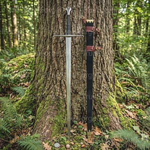 Puede incluir: Una espada larga y plateada con empuñadura y guarda negras, apoyada en un árbol. La espada tiene una hoja pulida y una empuñadura decorativa. Una vaina negra con detalles de cuero rojo descansa al lado, en un entorno forestal.