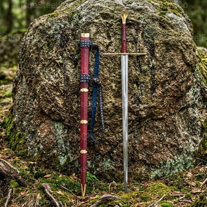 Peut inclure: Une épée médiévale avec une lame argentée et une poignée dorée est dressée à côté de son fourreau rouge. Le fourreau est orné d'accents dorés et d'une sangle bleue. L'épée et le fourreau sont posés contre un grand rocher dans un cadre forestier.