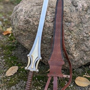 May include: A double-edged sword with a silver blade, a gold-toned metal crossguard featuring curved accents, and a brown leather-wrapped grip. Beside the sword is a matching brown leather scabbard with embossed detailing and leather straps.