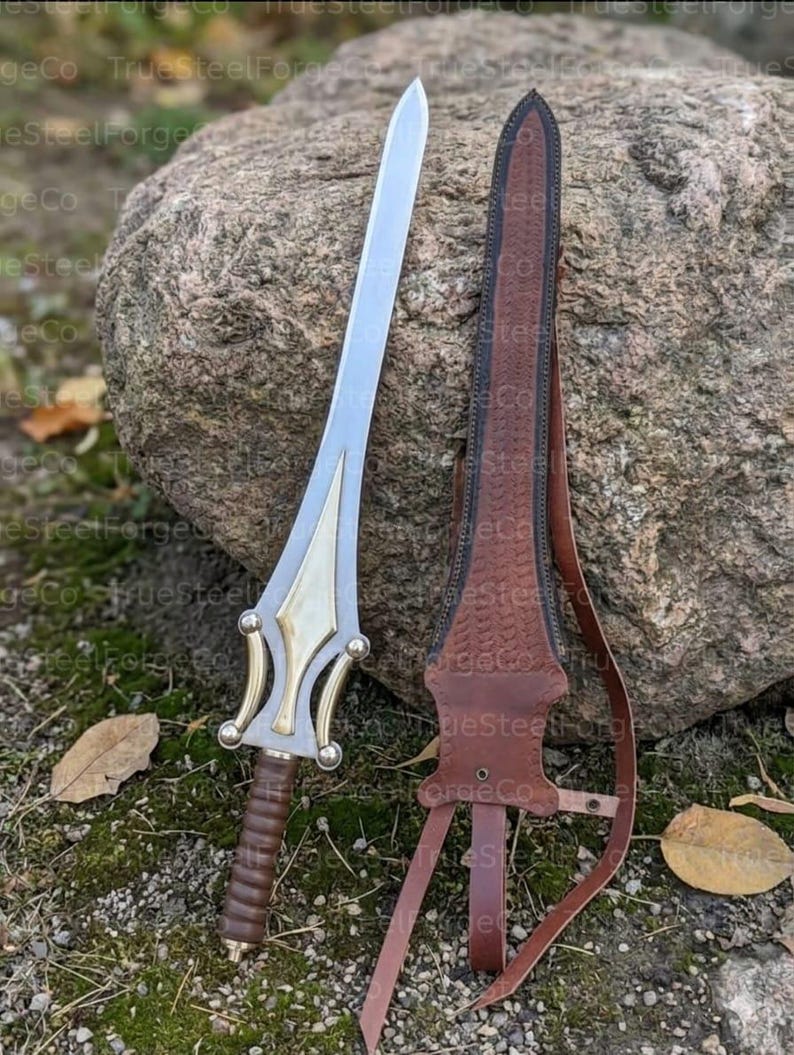 May include: A double-edged sword with a silver blade, a gold-toned metal crossguard featuring curved accents, and a brown leather-wrapped grip. Beside the sword is a matching brown leather scabbard with embossed detailing and leather straps.