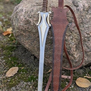 May include: A silver-toned metal sword with a brown ribbed handle and a decorative gold-colored crossguard. Beside the sword is a brown leather sheath featuring a textured herringbone pattern and a long leather strap for carrying or attachment.