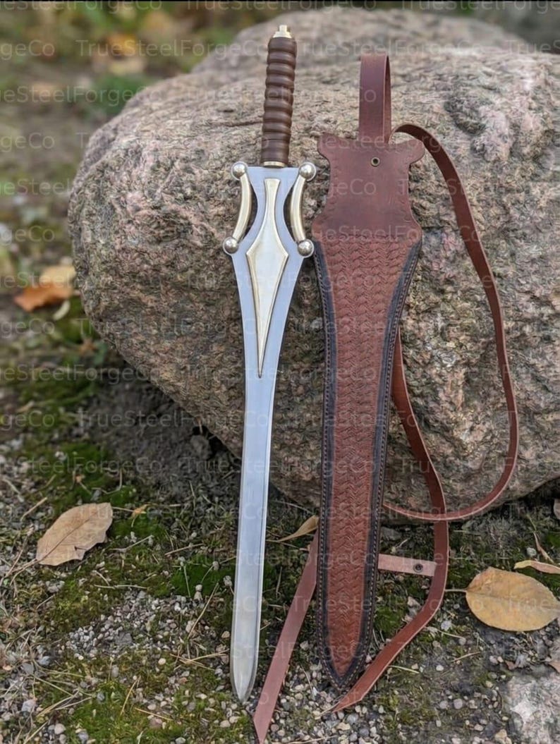 May include: A silver-toned metal sword with a brown ribbed handle and a decorative gold-colored crossguard. Beside the sword is a brown leather sheath featuring a textured herringbone pattern and a long leather strap for carrying or attachment.