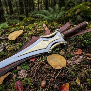 May include: A double-edged steel dagger featuring a polished brass crossguard and a brown leather-wrapped grip. The dagger rests inside a brown leather sheath with visible stitching and a leather strap, showcasing a metallic finish and brass accents.