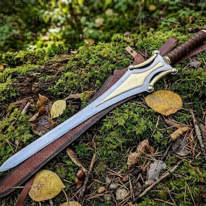 May include: A double-edged sword with a silver-toned blade and a gold-toned metal crossguard featuring curved accents. The handle is wrapped in brown leather, and the sword rests partially inside a brown leather scabbard with a strap.
