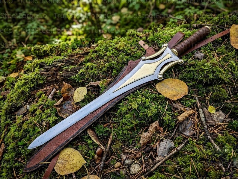 May include: A double-edged sword with a silver-toned blade and a gold-toned metal crossguard featuring curved accents. The handle is wrapped in brown leather, and the sword rests partially inside a brown leather scabbard with a strap.
