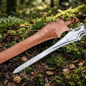 May include: A silver-colored metal sword with a pointed blade, a decorative crossguard featuring circular accents, and a black ribbed handle. The sword is accompanied by a tan leather scabbard with a strap and buckle closure mechanism.