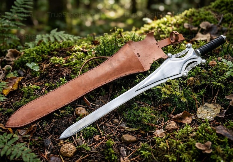 May include: A silver-colored metal sword with a pointed blade, a decorative crossguard featuring circular accents, and a black ribbed handle. The sword is accompanied by a tan leather scabbard with a strap and buckle closure mechanism.