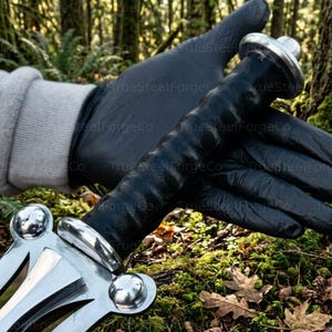 May include: A sword handle featuring a black textured grip, a polished silver-colored metal crossguard with three rounded decorative accents, and a matching silver-colored pommel at the end of the handle, held against a natural forest background.
