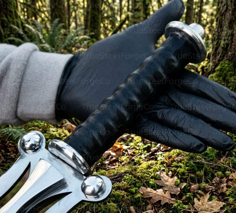 May include: A sword handle featuring a black textured grip, a polished silver-colored metal crossguard with three rounded decorative accents, and a matching silver-colored pommel at the end of the handle, held against a natural forest background.