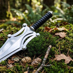 May include: A silver-colored metal dagger featuring a wide, pointed blade with cut-out sections and four rounded decorative studs. The handle is black with a ribbed, cylindrical grip and a metallic pommel at the end of the hilt.