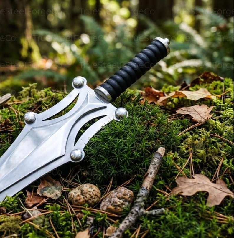 May include: A silver-colored metal dagger featuring a wide, pointed blade with cut-out sections and four rounded decorative studs. The handle is black with a ribbed, cylindrical grip and a metallic pommel at the end of the hilt.