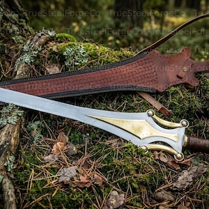 May include: A silver-toned metal sword featuring a polished brass inlay on the blade near the hilt, a brown leather-wrapped handle, and a matching brown leather scabbard with intricate embossed patterns and a long strap for carrying.