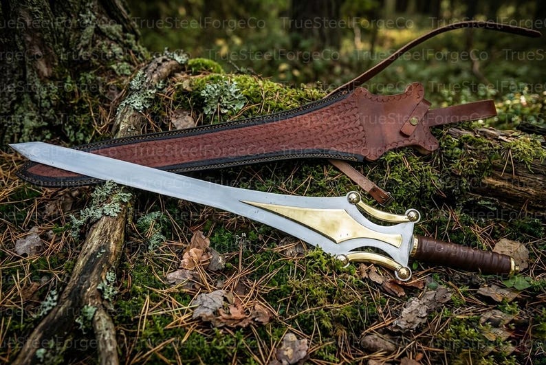 May include: A silver-toned metal sword featuring a polished brass inlay on the blade near the hilt, a brown leather-wrapped handle, and a matching brown leather scabbard with intricate embossed patterns and a long strap for carrying.
