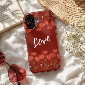 Könnte beinhalten: Eine rote Handyhülle mit einem floralen Mohnblumenmuster und dem Wort "Love" in weißer Schrift. Die Hülle hat eine glänzende Oberfläche und einen Kameraausschnitt. Die Mohnblumen blühen in voller Pracht.