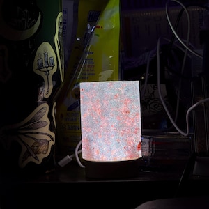 May include: A rectangular, illuminated lamp with a glittery, speckled surface. The lamp emits a soft, warm glow, with red and silver accents. The base is a warm, light brown color. The lamp is plugged in with a white cord.