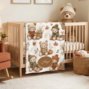 May include: A white baby blanket featuring a repeating pattern of teddy bears wearing knit hats and scarves, pumpkins, and autumn leaves. The blanket includes a central oval emblem with the name Hazel printed in a cursive script font.