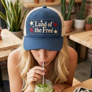 May include: A navy blue trucker hat with a beige mesh back and a rectangular patch on the front. The patch features the text Land of the Free in white lettering, surrounded by small red and white stars on a dark blue background.