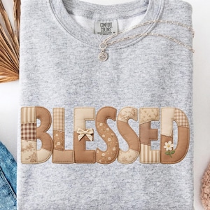 Peut inclure: Un sweat-shirt gris clair avec le mot "BLESSED" appliqué en tissu patchwork. Les lettres sont de différentes nuances de marron et de beige, avec un petit nœud et un détail floral. Un collier argenté et une paire de jeans sont également visibles.