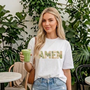 Könnte beinhalten: Ein weißes T-Shirt mit dem Wort "AMEN" in einem Patchwork-Design aus grünem und beigem Stoff. Die Person trägt Jeansshorts und hält ein grünes Getränk. Der Hintergrund zeigt üppige grüne Pflanzen und Cafétische.