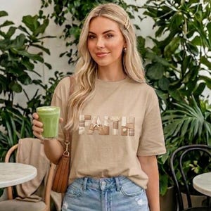 Könnte beinhalten: Ein beiges T-Shirt mit dem Wort "FAITH" in einem Patchwork-Design. Die Person trägt Jeansshorts und hält ein grünes Getränk. Der Hintergrund zeigt üppige grüne Pflanzen und Cafétische.