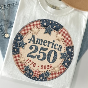 May include: A white t-shirt featuring a circular graphic with a patchwork design of red and white gingham and blue star-patterned fabric. The centre text reads America 250 with the years 1776 and 2026 printed below in a dark blue font.