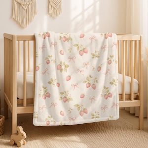 Könnte beinhalten: Eine cremefarbene Babydecke mit einem wiederkehrenden Muster aus roten Erdbeeren, grünen Blättern und kleinen rosa Schleifen. Die Decke liegt über einem hellen Holzbettchen und zeigt eine weiche Textur sowie einen sauber verarbeiteten Rand.