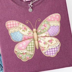 May include: A purple sweatshirt with a large, appliqued butterfly design. The butterfly features patchwork wings with floral, polka dot, and gingham patterns, accented with lace trim. A silver necklace with a small pendant is visible.