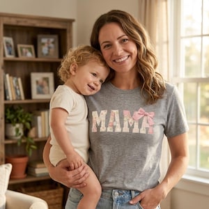 Peut inclure: Un t-shirt gris avec le mot "MAMA" en motifs floraux et à carreaux, et un nœud rose. Une femme tient un bébé en barboteuse blanche. Une bibliothèque en bois est en arrière-plan.