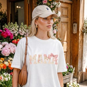 May include: A white t-shirt with the word "MAMA" in floral and plaid patterns, topped with a pink bow. The person is wearing a beige baseball cap with a navy blue logo and gold hoop earrings. A brown shoulder bag is visible.