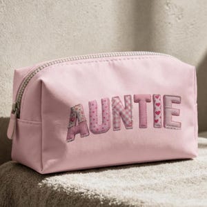 May include: A pink cosmetic bag with the word "AUNTIE" embroidered across the front. The letters are in various pink patterns, including floral, polka dots, and hearts. The bag has a silver zipper.