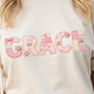 May include: A cream-coloured sweatshirt with the name "GRACE" appliquéd across the front. The letters are patchwork style, featuring pink floral, polka dot, and gingham patterns, with small rose accents and bows.