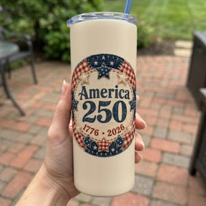 May include: A cream-coloured cylindrical tumbler with a clear lid and a blue straw. The front features a circular graphic with a red, white, and blue plaid border, stars, and the text America 250 with the years 1776 and 2026 printed in blue and red.