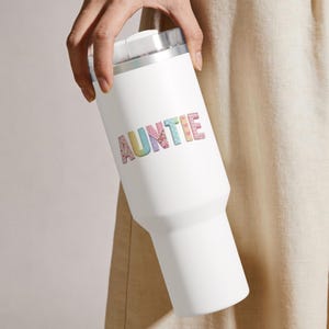 May include: A white insulated tumbler with a silver rim and lid. The word "AUNTIE" is displayed in colorful, patterned letters across the front. The tumbler is designed to keep beverages cold or hot.