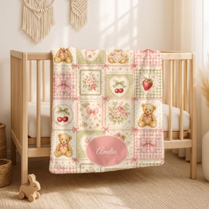 Puede incluir: Una manta suave para bebé con diseño de patchwork de cuadros vichy en rosa claro y verde, ositos de peluche, lazos rosas, ramos de flores, cerezas y una fresa. Un óvalo rosa central contiene el texto en cursiva blanca Amelia.