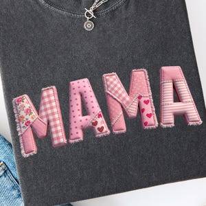 May include: Dark grey t-shirt featuring the word "MAMA" in pink patchwork letters. The letters are decorated with floral, gingham, polka dot, heart, and striped patterns. A silver necklace with a pendant is also visible.