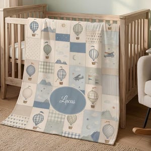 May include: A soft baby blanket featuring a patchwork design in shades of blue, cream, and gray. The pattern includes illustrations of hot air balloons, airplanes, mountains, stars, and clouds. A central blue oval contains the name Lucas in white.
