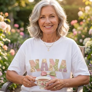 May include: A woman wearing a white t-shirt with the word "NANA" in patchwork letters and a bow design. She is holding a floral teacup and saucer. The woman is smiling and sitting outside in a garden.