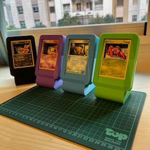 Op de afbeelding: Vier 3D-geprinte displaystandaards in zwart, paars, lichtblauw en limoengroen, elk met een andere Pokémon-ruilkaart. De kaarten bevatten illustraties van Axew, Abra, Feebas en Paras, met zichtbare tekst en spelstatistieken.