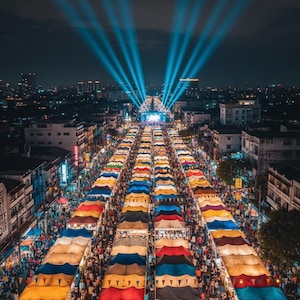 May include: Aerial view of a night market with rows of colorful tents and crowds of people. Bright blue spotlights shine down the center of the market, illuminating the scene against the dark sky and city skyline.