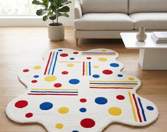 Op de afbeelding: Een wit, onregelmatig gevormd vloerkleed met een patroon van rode, blauwe en gele cirkels en strepen. Het vloerkleed ligt op een houten vloer in een woonkamer. Het ontwerp is modern en speels.