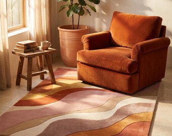 Op de afbeelding: Een rechthoekig vloerkleed met een golvend, veelkleurig patroon in tinten terracotta, oudroze, crème en mosterdgeel. Het kleed heeft een zachte, pluizige textuur en ligt op een lichte vloer onder een fauteuil van fluweel.