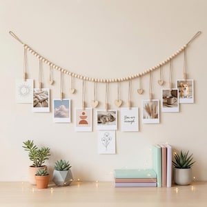 Könnte beinhalten: Eine dekorative Fotowand mit einer Holzperlen-Girlande und hängenden Fotos im Polaroid-Stil. Die Fotos werden von Holzklammern und herzförmigen Akzenten gehalten. Die Fotos zeigen Bilder einer Katze, eines Hundes und andere minimalistische Designs. Kleine Topfpflanzen und Bücher sind auf der Oberfläche.