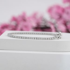 Op de afbeelding: Een delicate zilveren tennisarmband met talrijke kleine, ronde, heldere edelstenen. De armband wordt getoond op een wit oppervlak, met een vage roze bloemenachtergrond.