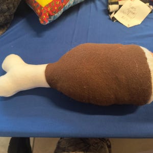 May include: A plush dog toy shaped like a turkey leg. The toy features a brown drumstick, a white bone, and a colorful end. The toy is designed to resemble a cooked turkey leg.